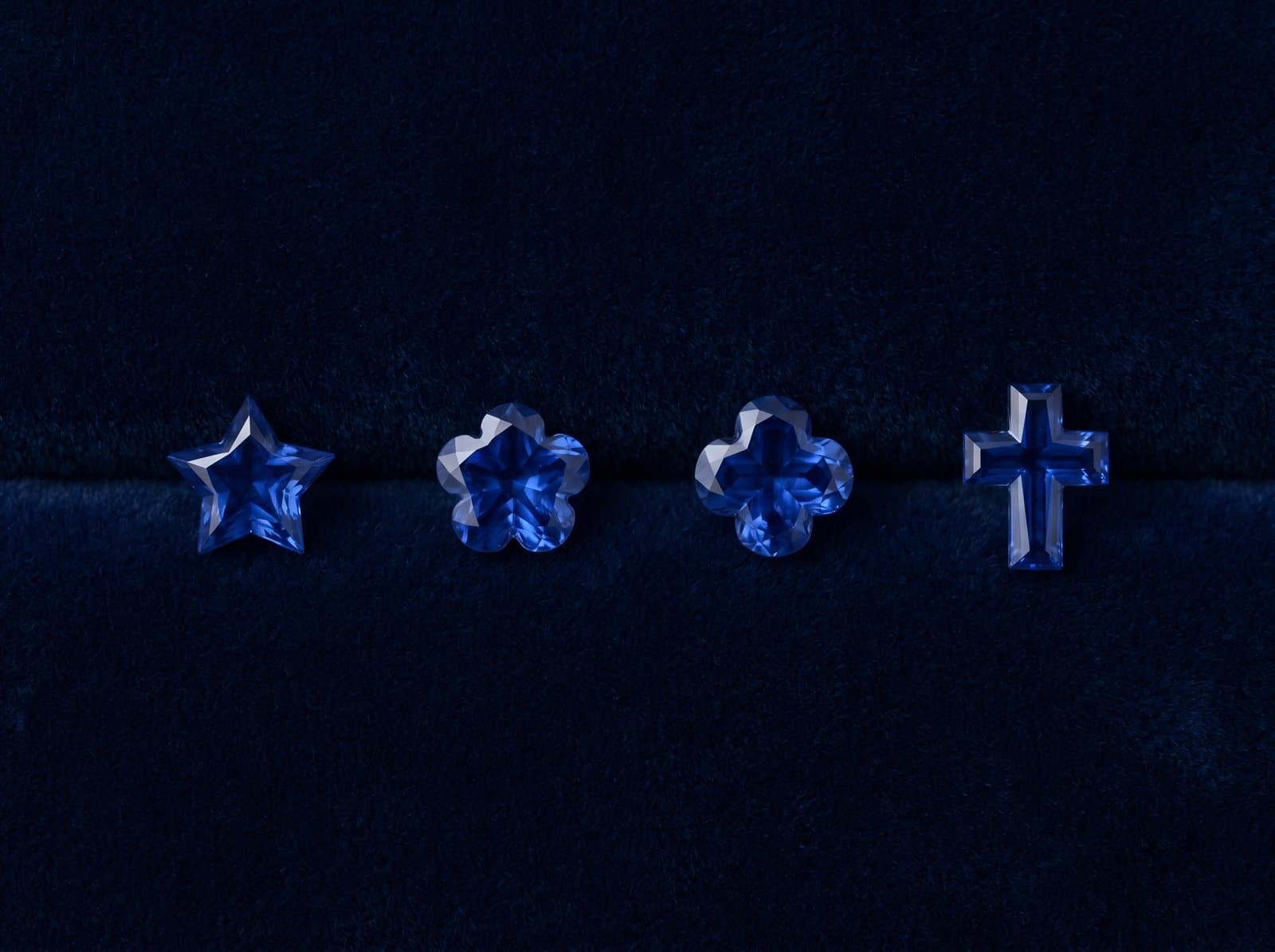 Custom-cut blue sapphires in fancy shapes — star, flower, clover, and cross — arranged on a deep navy velvet surface